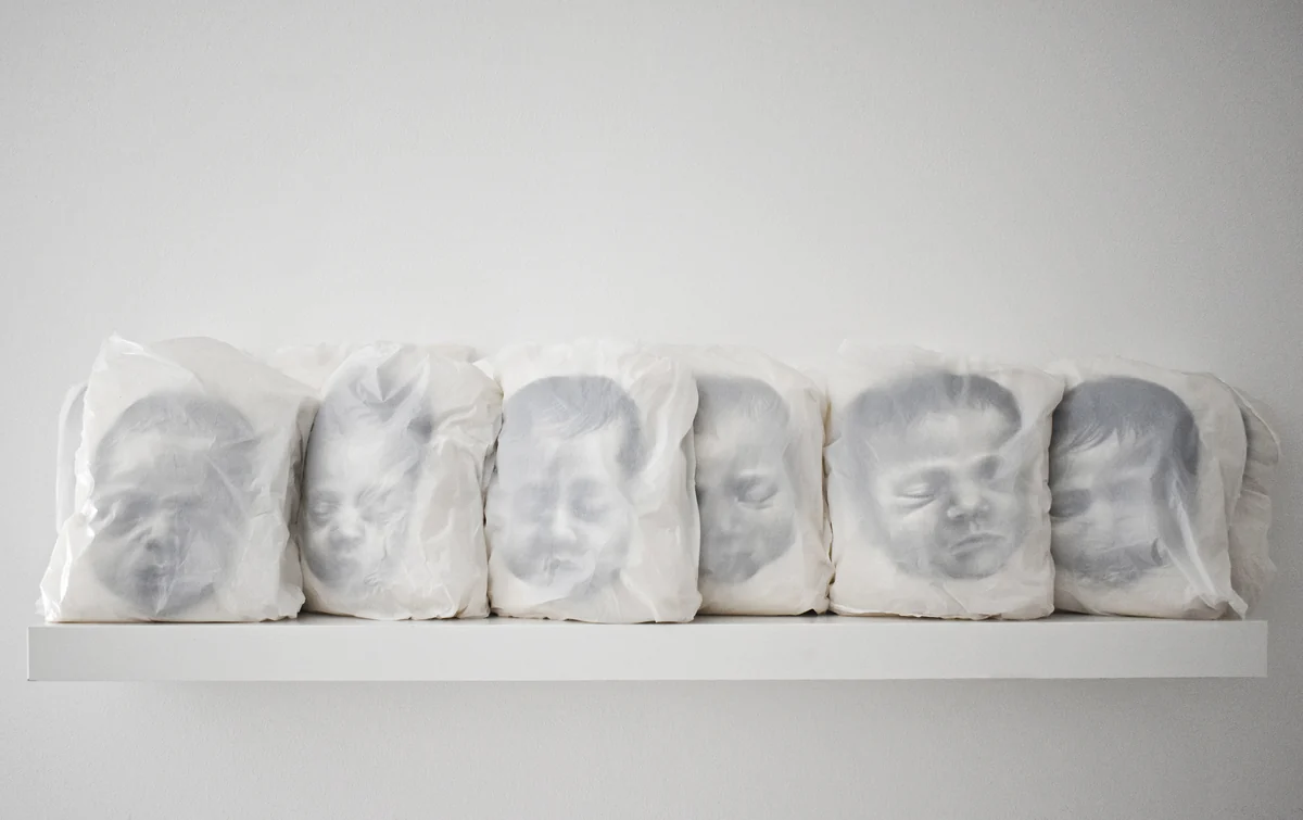 Angels of Our Time, a row of pale covered faces on a white shelf